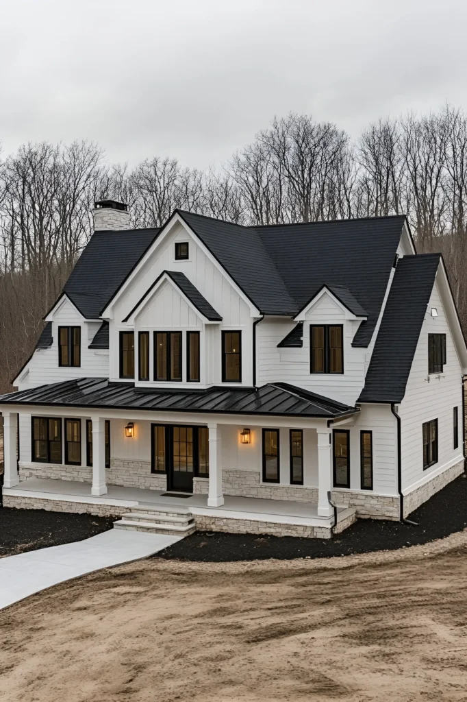 Modern white farmhouse with black window frames, stone foundation, board-and-batten siding, and a wooded background.