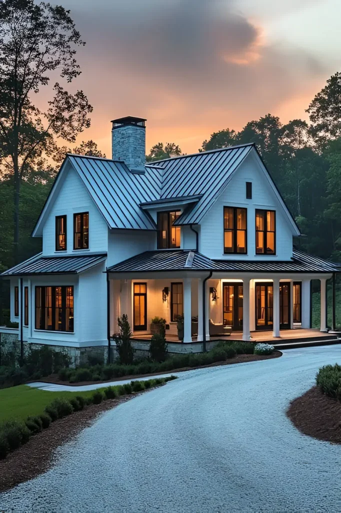 Modern white farmhouse with a metal roof, stone chimney, wraparound porch, and warm interior lighting, set against a twilight sky.