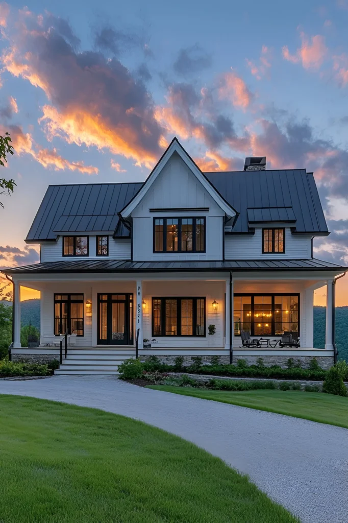 Modern white farmhouse with a black metal roof, large windows, wraparound porch, and a vibrant sunset sky in the background.