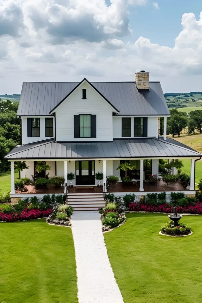 Modern white farmhouse with a black metal roof, stone chimney, wraparound porch, vibrant flower gardens, and a white stone pathway.