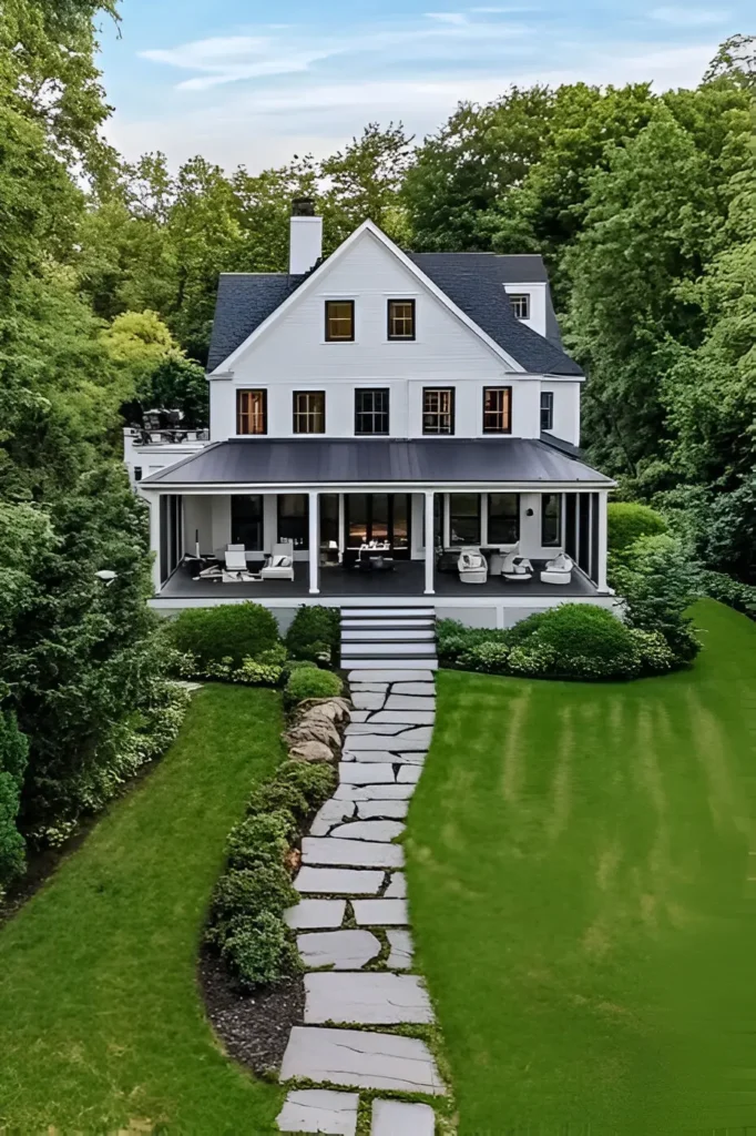 Modern white farmhouse with a black roof, wraparound porch, stone pathway, and lush green backyard surrounded by trees.