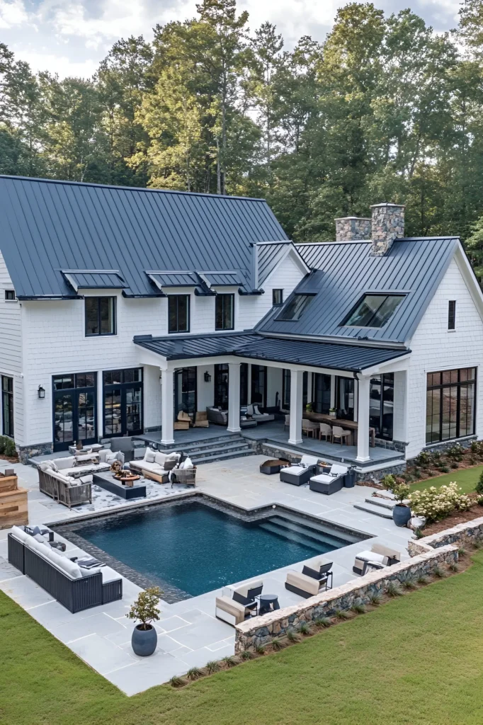 Modern white farmhouse with a black metal roof, resort-style pool, outdoor seating areas, and stone accents surrounded by trees.