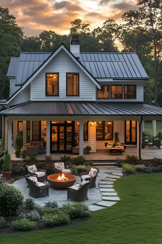 A modern white farmhouse with a stylish patio, fire pit surrounded by wicker chairs, and glowing interiors, set against a scenic sunset.