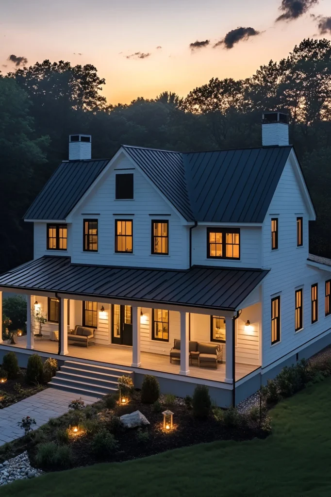 Modern white farmhouse with a wraparound porch, illuminated landscaping, and a peaceful evening backdrop.