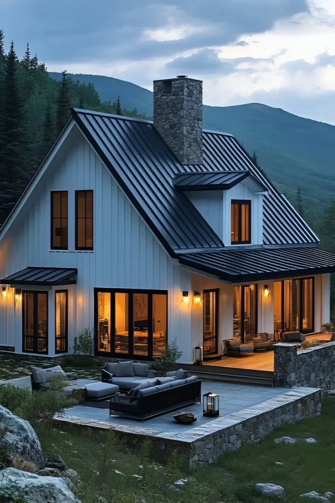 Modern white farmhouse with a stone chimney, black-framed windows, and a scenic mountain backdrop illuminated by warm interior lights.