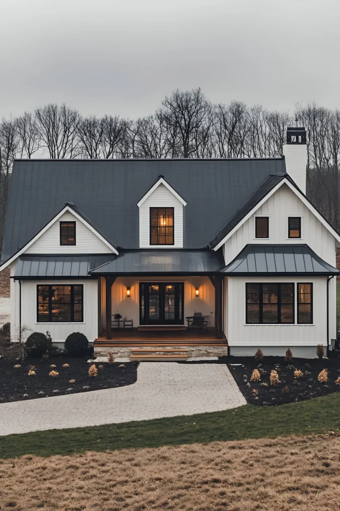 Modern white farmhouse with gabled roof, black windows, and warm porch lighting set against a wintery woodland backdrop.