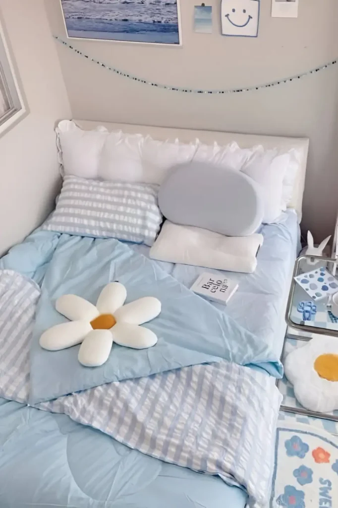 A cozy small bedroom featuring soft blue bedding, striped pillows, and a daisy-shaped pillow. The decor includes coastal wall art, a pastel aesthetic, and a calming, playful atmosphere perfect for a minimalist or teen bedroom.