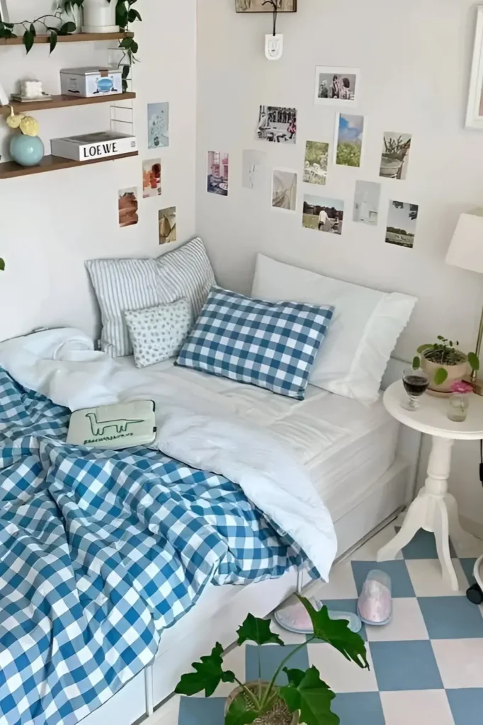 A small bedroom featuring blue gingham bedding, striped pillows, photo wall decor, and a checkerboard floor. The space includes a side table with plants, creating a cozy, modern, and refreshing atmosphere.