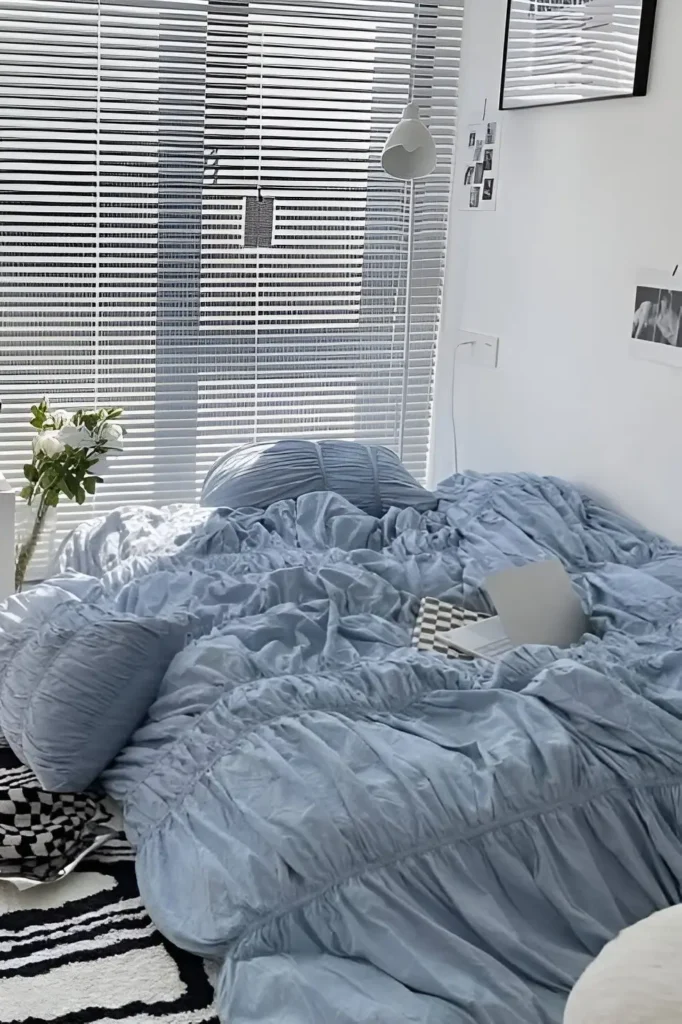 Modern minimalist bedroom featuring soft blue ruched bedding, a white lamp, large blinds, fresh flowers, and black-and-white rug accents.