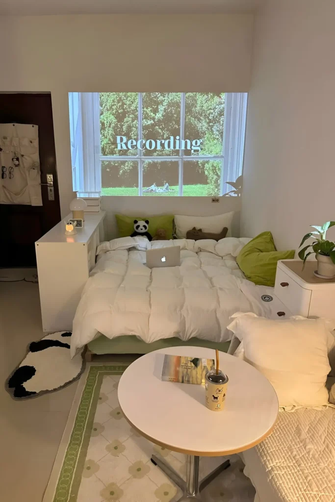 Cozy small bedroom with white bedding, green accent pillows, a projection wall displaying visuals, and a small round table with a book and drink. Decor includes plants and soft lighting for a relaxing vibe.