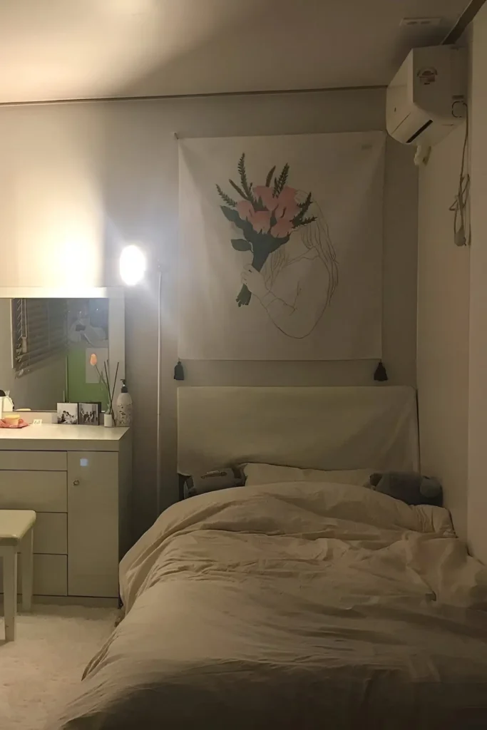 Small minimalist bedroom with beige bedding, soft lighting, floral wall art, and a simple vanity setup for a calm and cozy atmosphere.