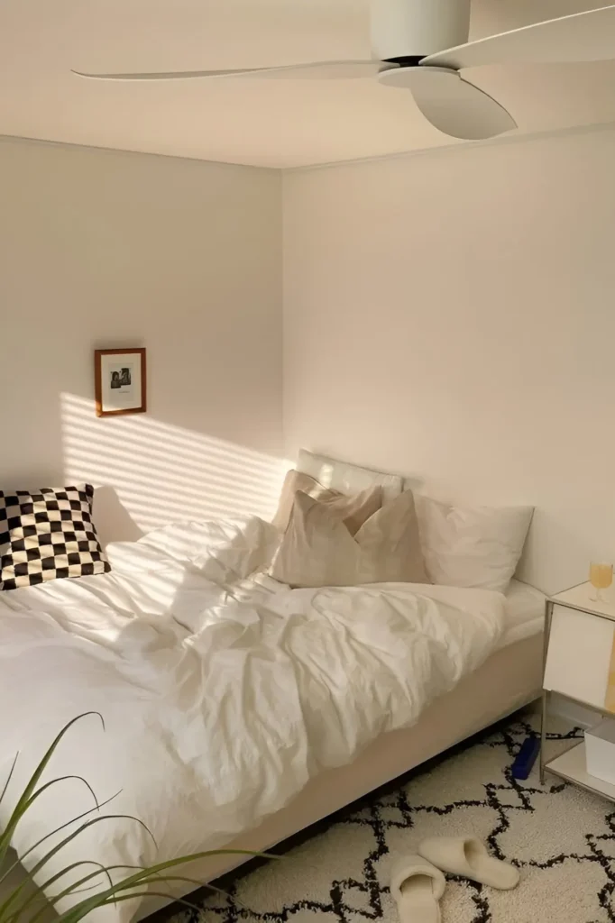 Minimalist bedroom with soft sunlight, neutral bedding, beige pillows, checkered accent cushion, and a serene, clutter-free design.