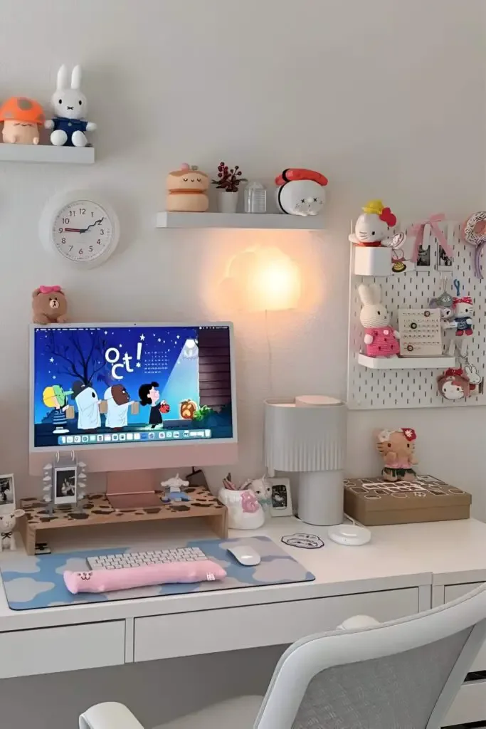 Kawaii-inspired desk setup with a pastel pink monitor, cloud-shaped light, plush toys, decorative shelves, and a soft wrist rest on a patterned desk mat.