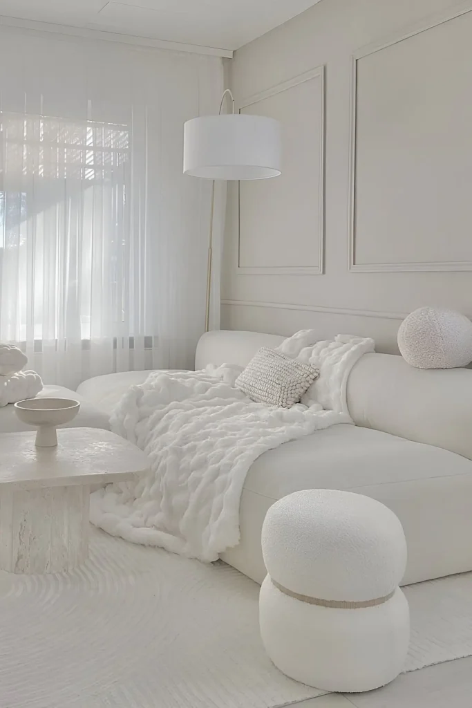 Cozy white living space featuring a curved sofa, fluffy throw, knit pillow, boucle ottoman, sculptural side table, and sheer curtains.