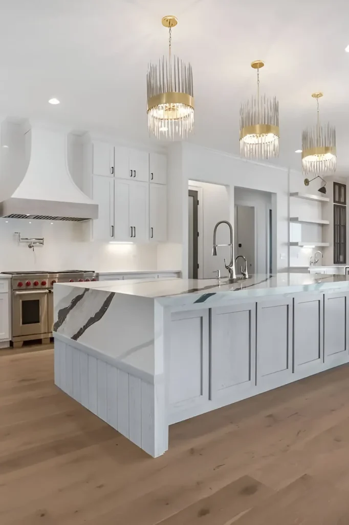 Modern white kitchen featuring a marble island, custom cabinetry, professional appliances, gold chandeliers, and wood floors.