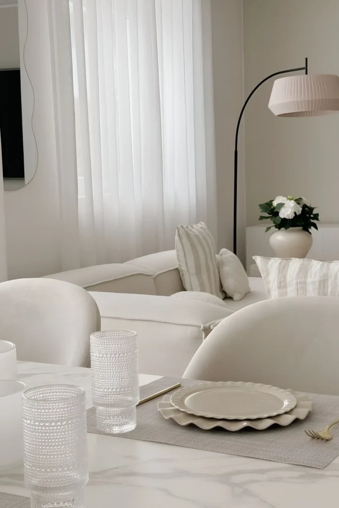 Minimalist dining and living room featuring a white table with textured glasses, wavy ceramic plates, and a cozy white sofa with striped cushions in the background, accented by a curved floor lamp.