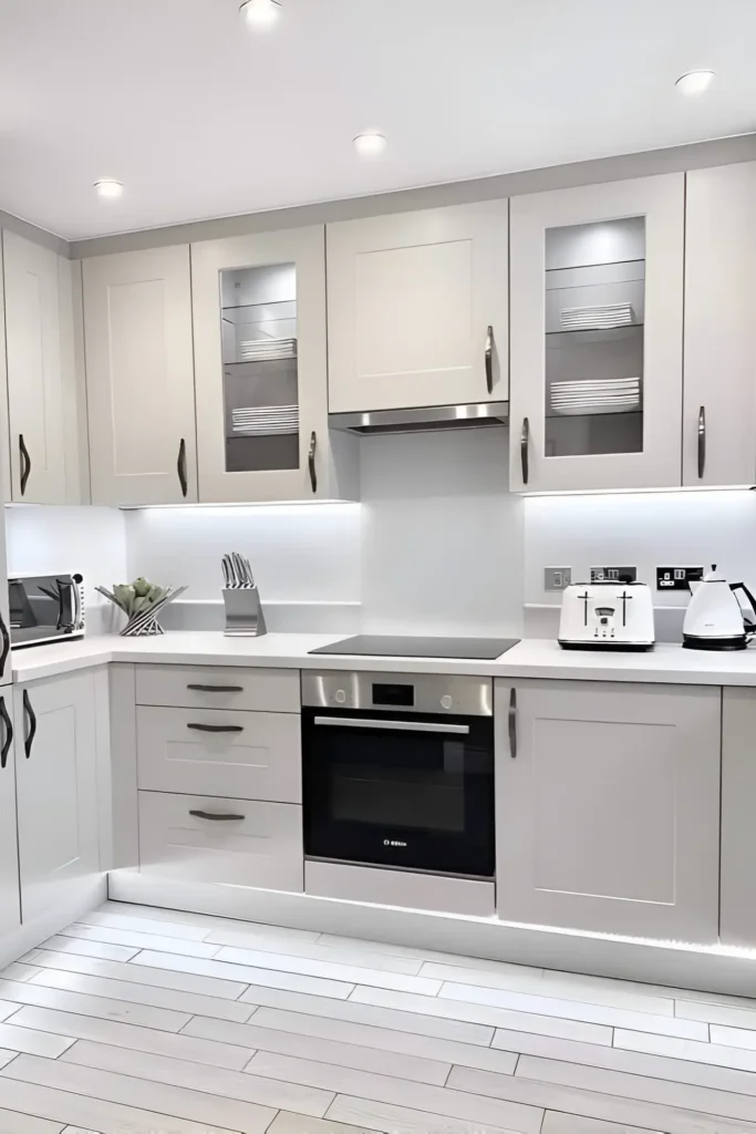 Compact white kitchen with glass-front cabinets, stainless steel appliances, and under-cabinet lighting.