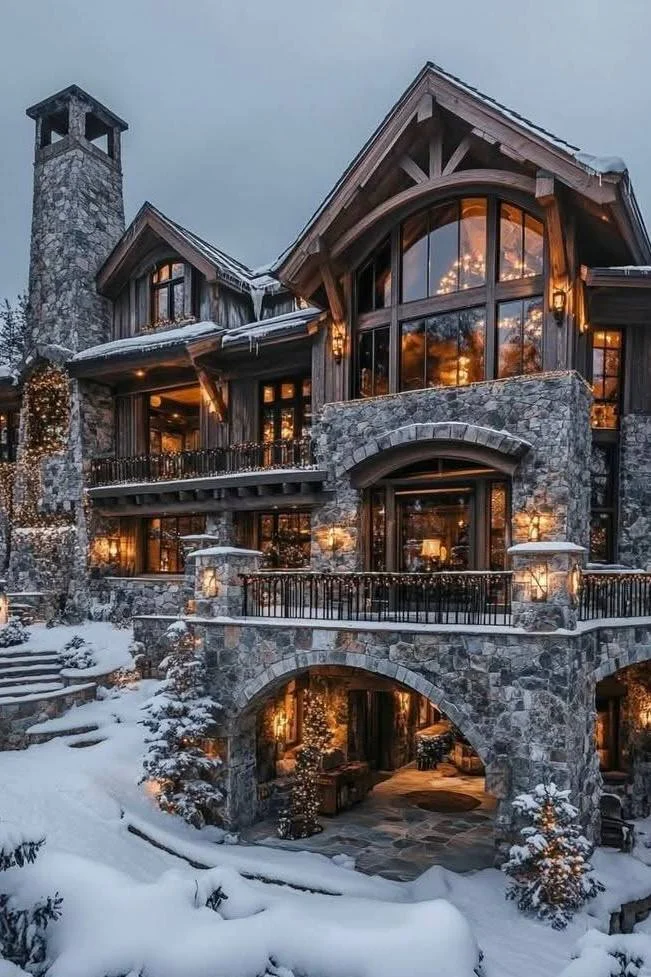 Luxury chalet with a grand stone archway entrance, multiple levels, balconies, and warm lighting, surrounded by snow.