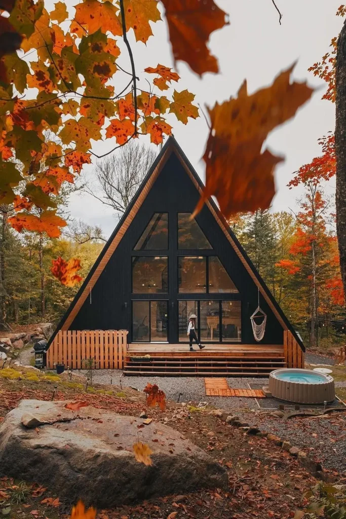 A black A-frame cabin with large glass windows, surrounded by autumn trees and a wooden deck with a hot tub in the foreground.