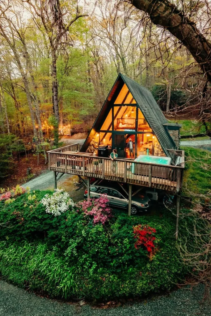 Elevated A-frame house surrounded by vibrant greenery and trees, featuring a spacious deck with a hot tub and illuminated interiors visible through large windows.