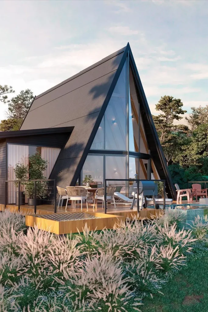 Modern black A-frame house with large glass windows, a furnished wooden deck, and surrounding greenery under a clear sky.