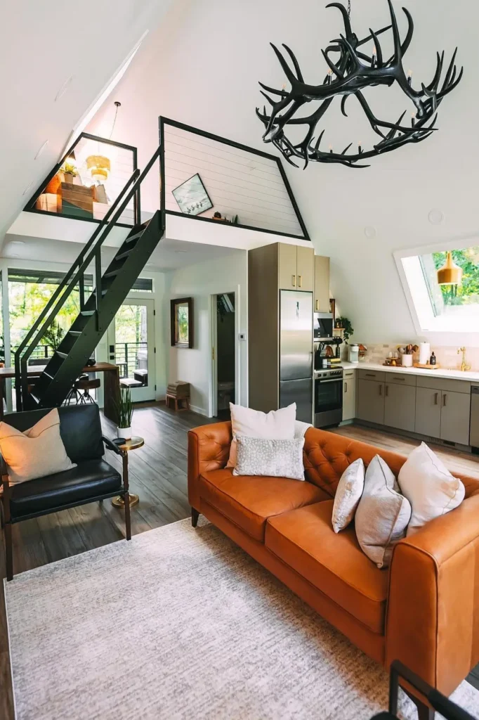 Interior of an A-frame house with a leather sofa, antler-style chandelier, loft area with black railings, and a modern kitchen in the background.