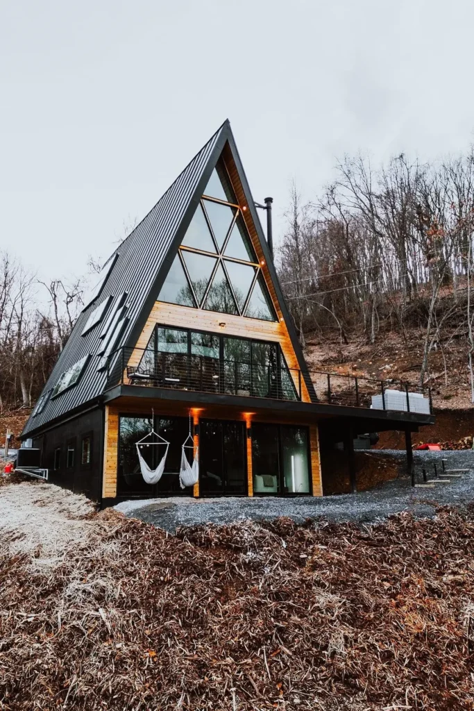 Large A-frame house with geometric glass windows, a wraparound deck, and hanging chairs, set on a wooded hillside with modern accents.