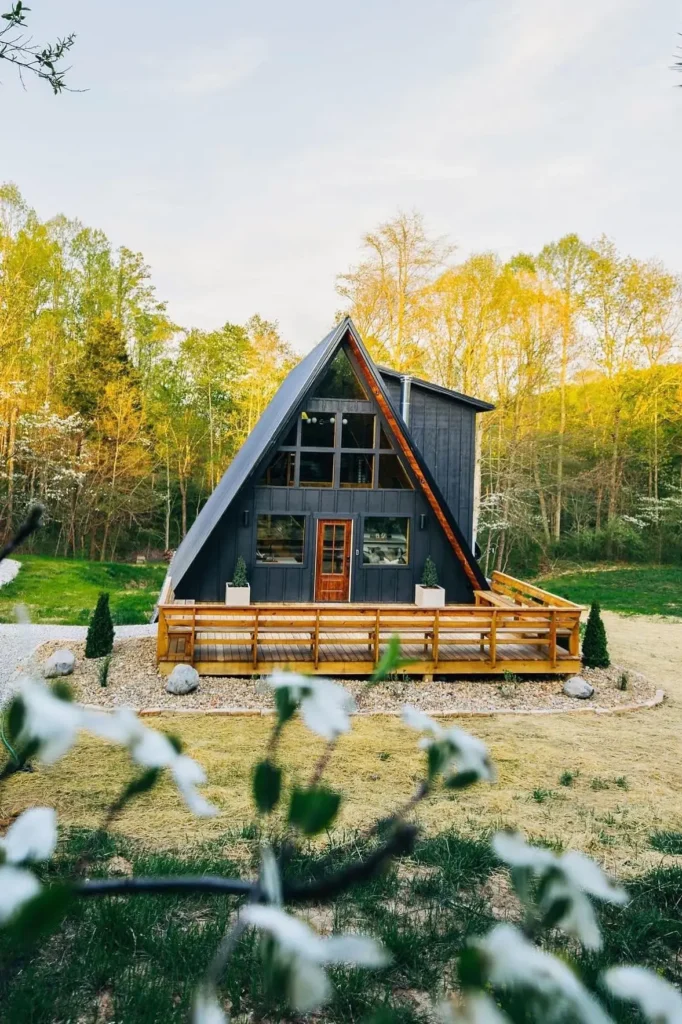 Dark-sided A-frame house with wooden trim, a spacious front deck, and a lush green backdrop on a sunny day.