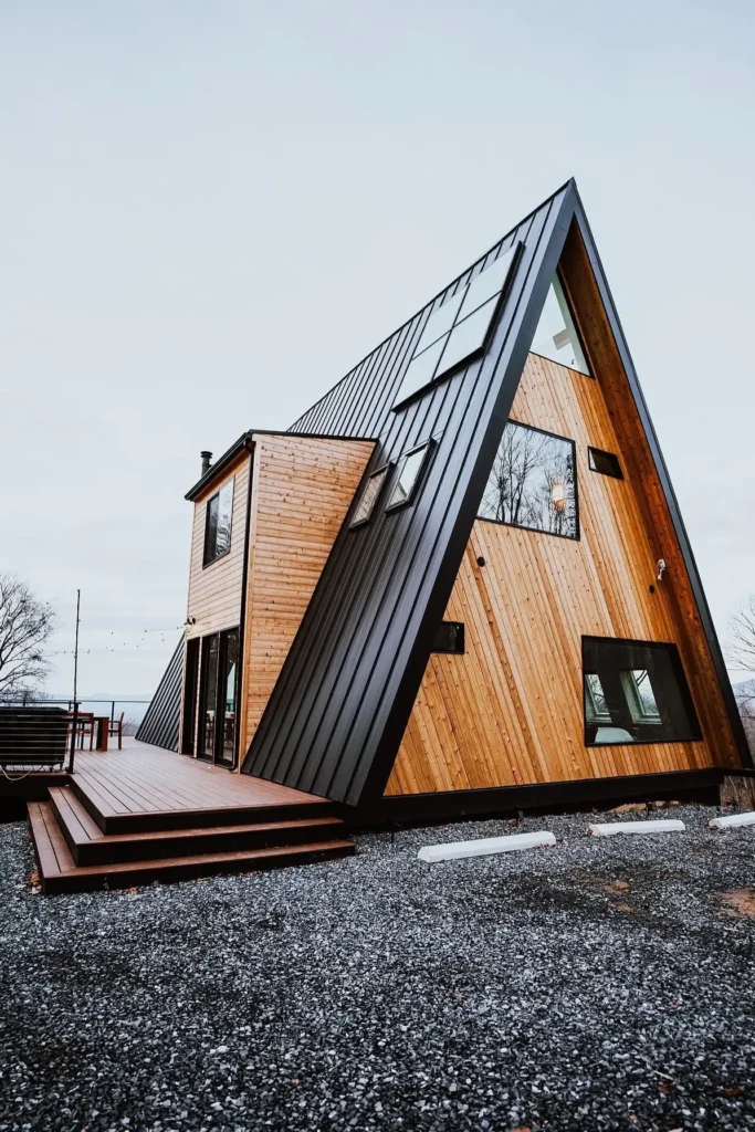 Contemporary A-frame house with black metal and wood siding, an extended deck, and large windows, set against an open sky and gravel pathway.