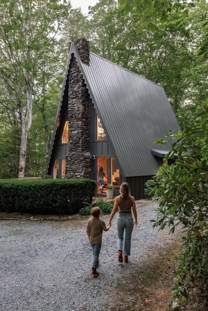 Rustic A-frame house with a tall stone chimney, surrounded by trees, with a mother and child walking toward the warmly lit entrance.