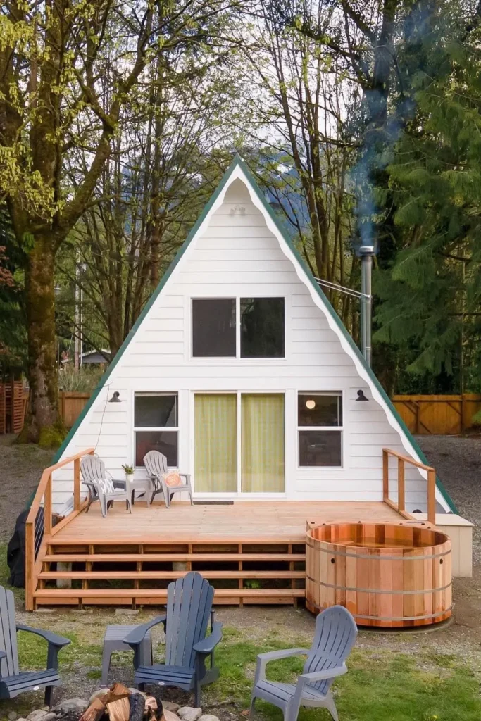 White A-frame house with a wooden deck, Adirondack chairs, a hot tub, and a backdrop of tall trees and greenery.