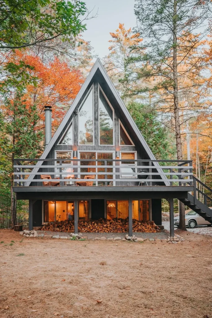 Black A-frame cabin with a spacious upper deck, large windows, and a woodpile below, surrounded by autumn-colored trees.