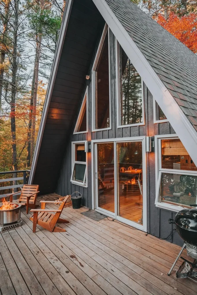 A-frame cabin deck with Adirondack chairs, a fire pit, large windows, and a view of autumn trees in the background.