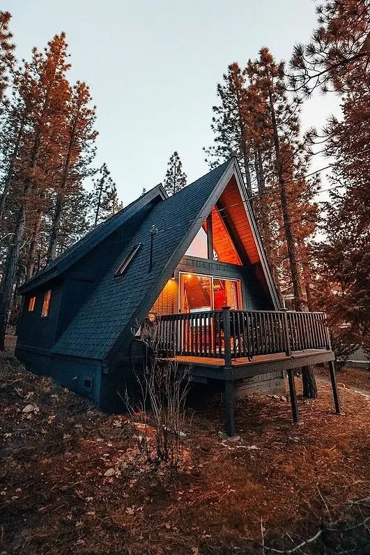 A-frame cabin with a dark exterior, warm interior lighting, and an elevated deck, surrounded by tall pine trees during twilight.