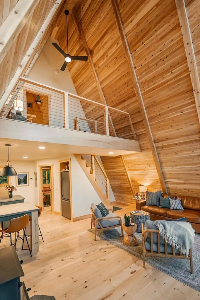 Interior of an A-frame house with natural wood walls, a loft, leather furniture, and a cozy, open-plan living and dining area.