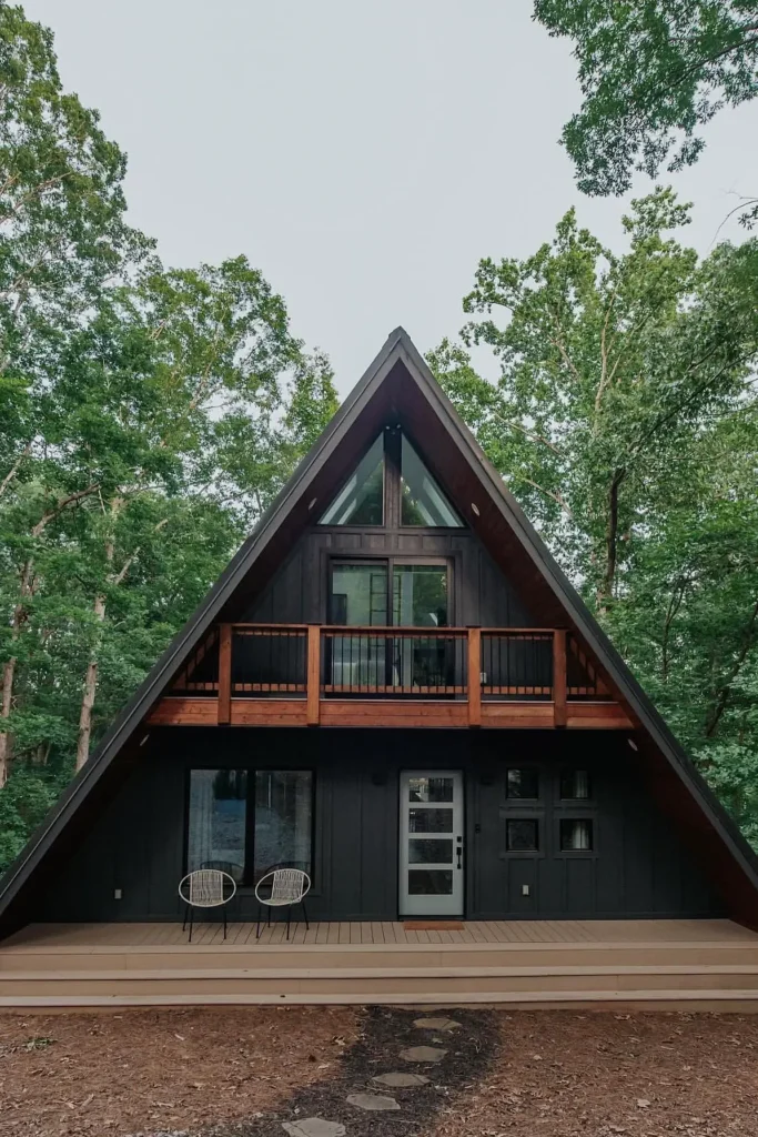 A-frame cabin with a dark exterior, an upper balcony, and a spacious lower deck surrounded by lush greenery.