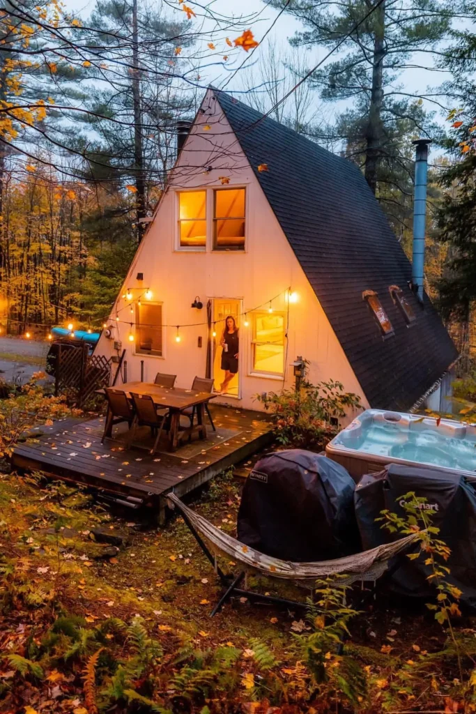 A-frame cabin with a deck, string lights, outdoor dining setup, a hot tub, and a hammock, surrounded by forest in the evening.