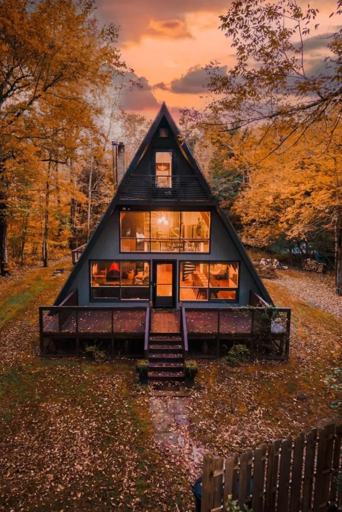 A-frame cabin with glowing windows, a spacious deck, and surrounded by vibrant autumn trees under a dramatic evening sky.