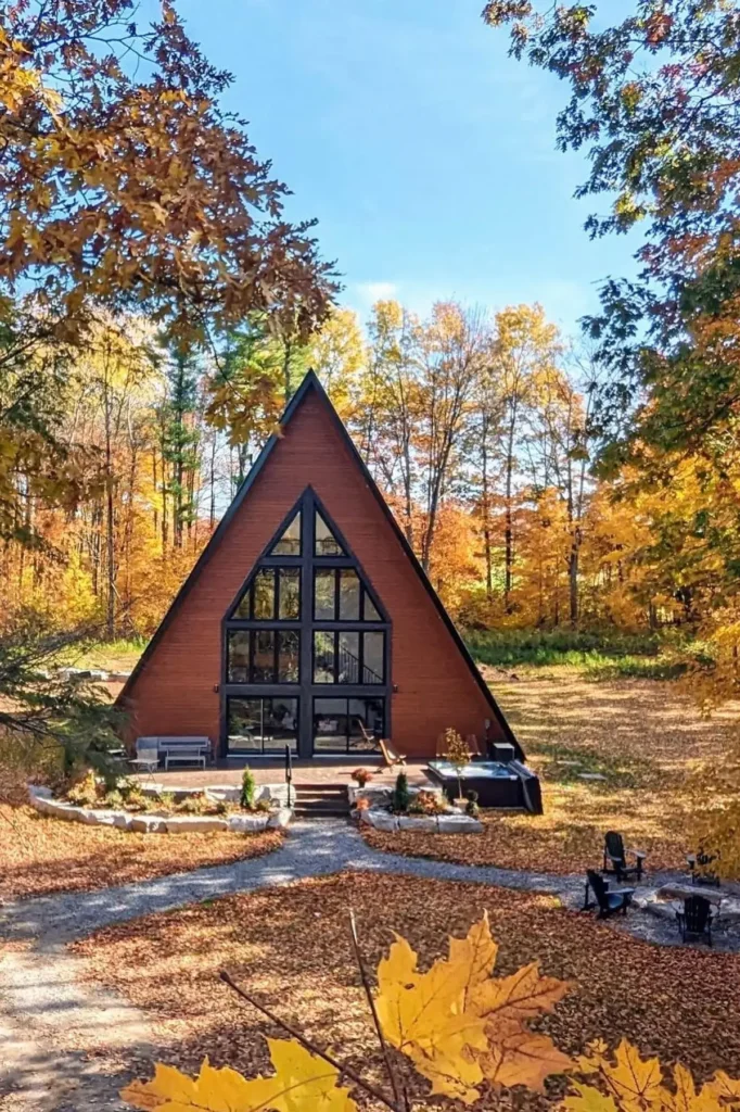 A-frame cabin with a wooden exterior, large glass windows, and an outdoor seating area, surrounded by vibrant autumn trees under a clear blue sky.