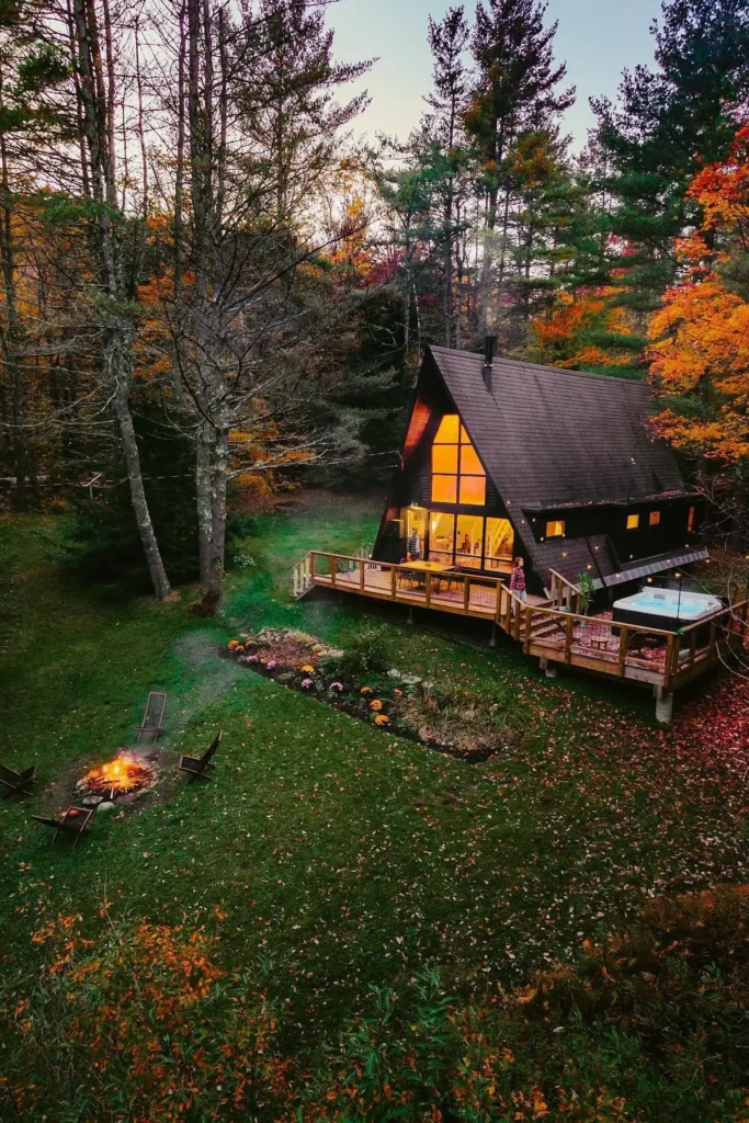 A-frame cabin with a wraparound deck, hot tub, fire pit, and vibrant autumn garden, surrounded by tall trees and lush greenery.