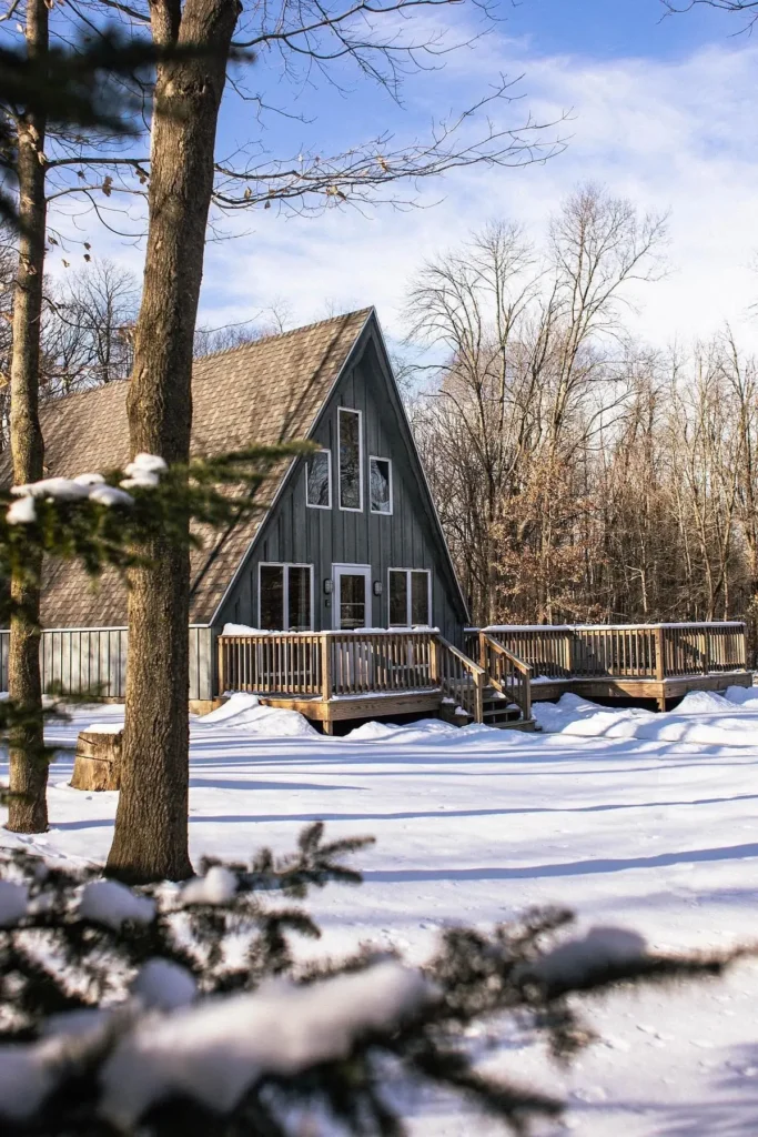 A-frame cabin with a spacious wooden deck, surrounded by snow-covered ground and bare winter trees under a bright blue sky.