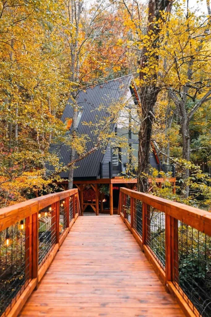 A-frame cabin surrounded by colorful autumn trees, with a wooden bridge leading to the entrance, creating a warm and inviting forest retreat.