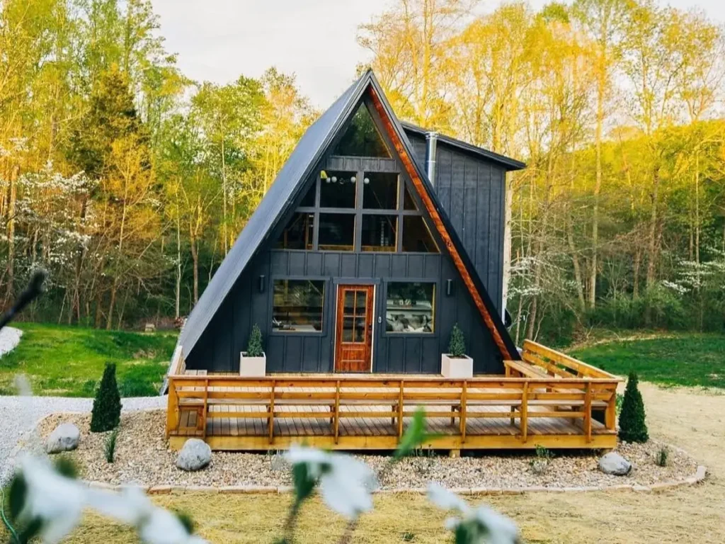 A-frame cabin with a wraparound deck, large windows, and modern planters, surrounded by vibrant greenery in a peaceful outdoor setting. Cozy A-Frame House