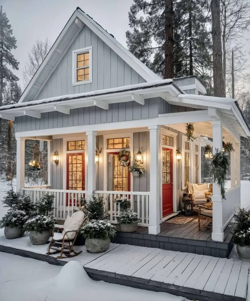 Gray cottage with red doors, festive garlands, frosted evergreens, and cozy porch seating.