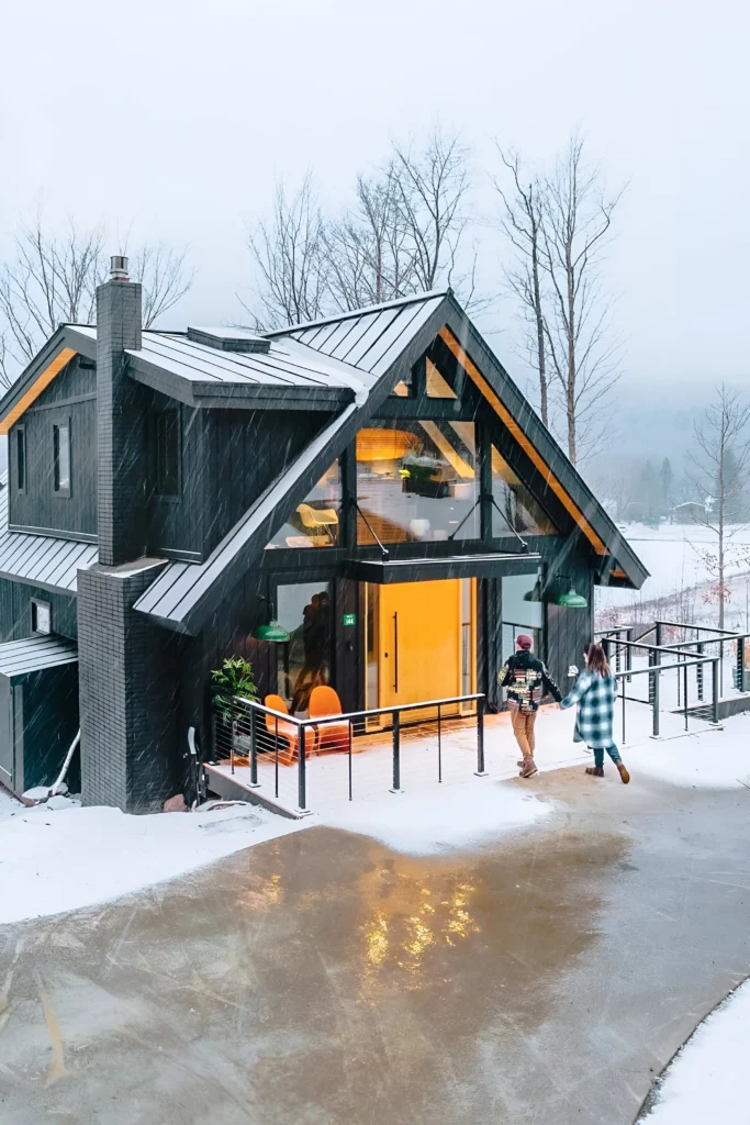 Modern A-frame cabin with large glass windows, dark exterior, and a snowy backdrop.