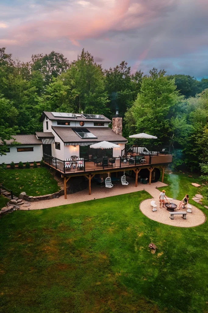 Modern sustainable cabin with solar panels, wraparound deck, and fire pit area surrounded by greenery.