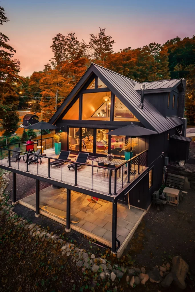 Contemporary A-frame cabin with a large balcony, lounge chairs, and glowing interiors against a sunset backdrop.