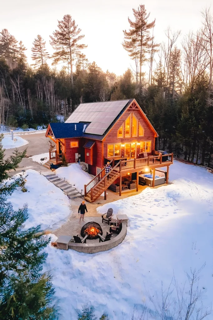 Sustainable log cabin with solar panels, fire pit, and snowy surroundings amidst a forest.