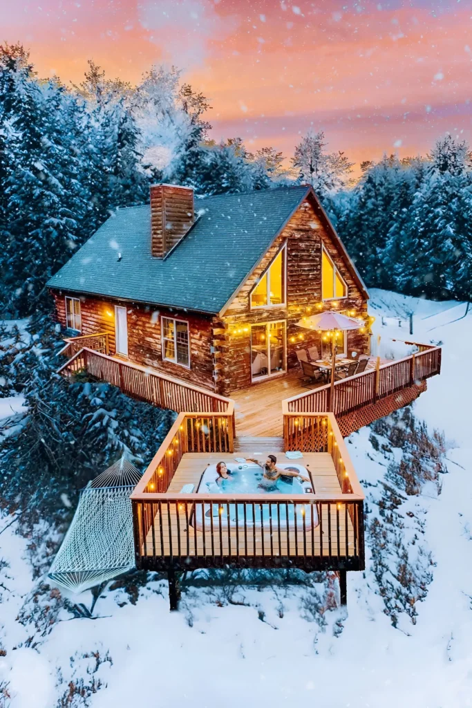 Winter cabin with a glowing deck, private hot tub, hammock, and snow-covered forest backdrop.