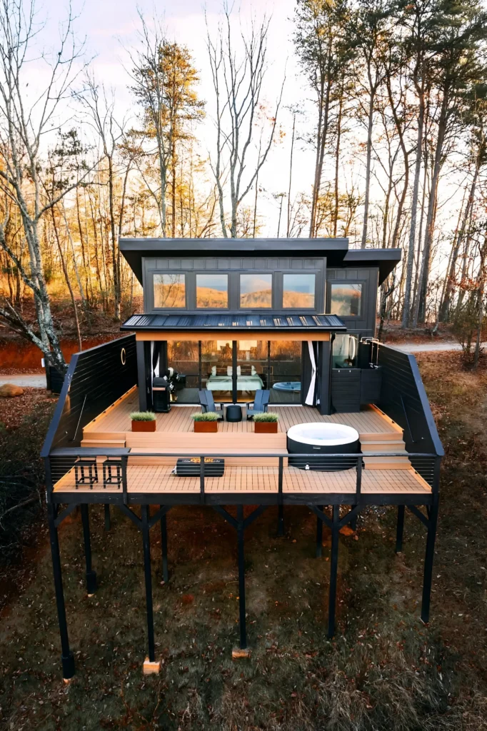 Modern elevated cabin with a large deck, hot tub, and floor-to-ceiling windows surrounded by a serene forest.